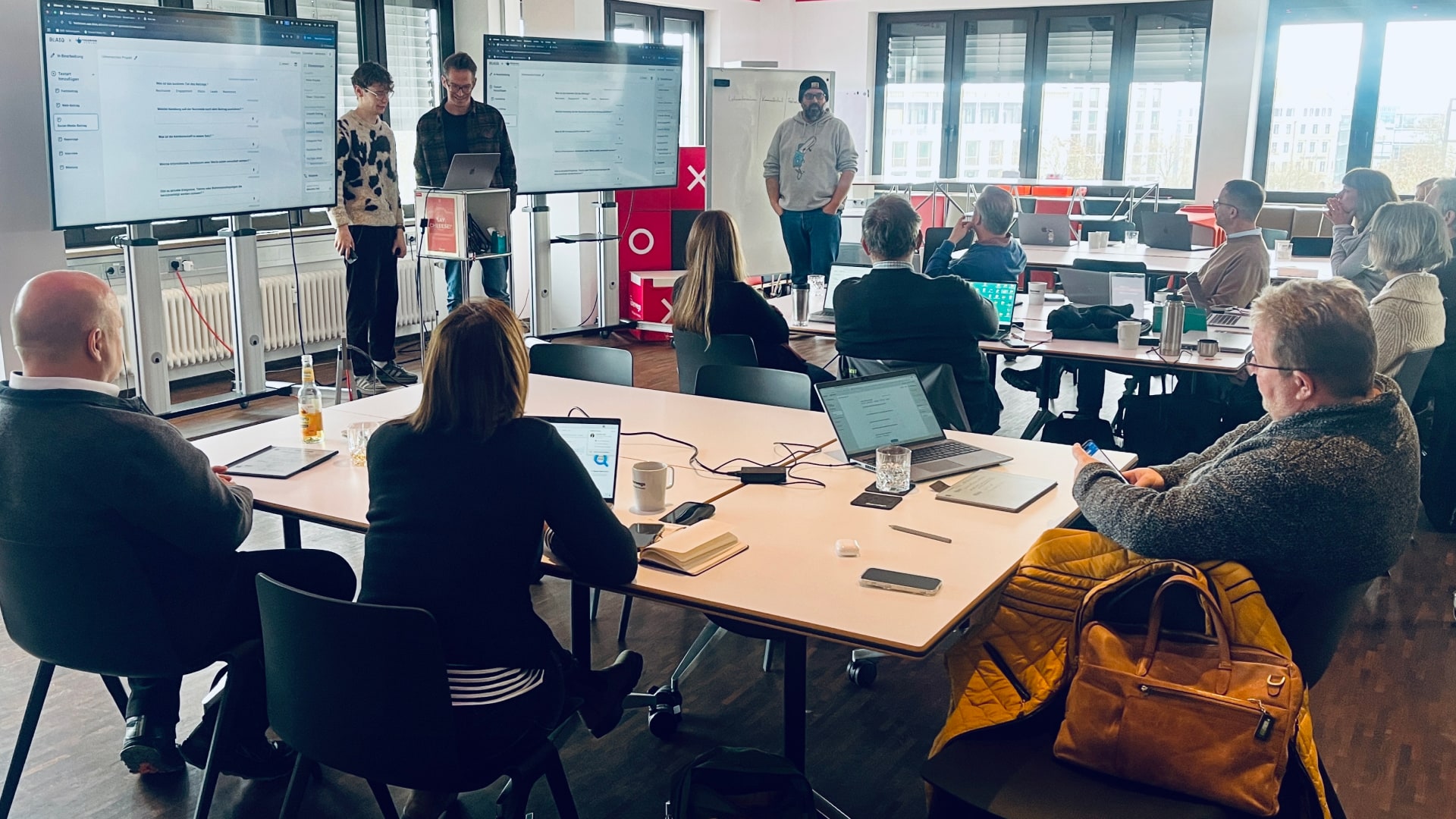 Workshop-Session mit rund zwölf Teilnehmenden an einem Konferenztisch; zwei Presenter zeigen die Blaiq-Plattform auf großen Monitoren